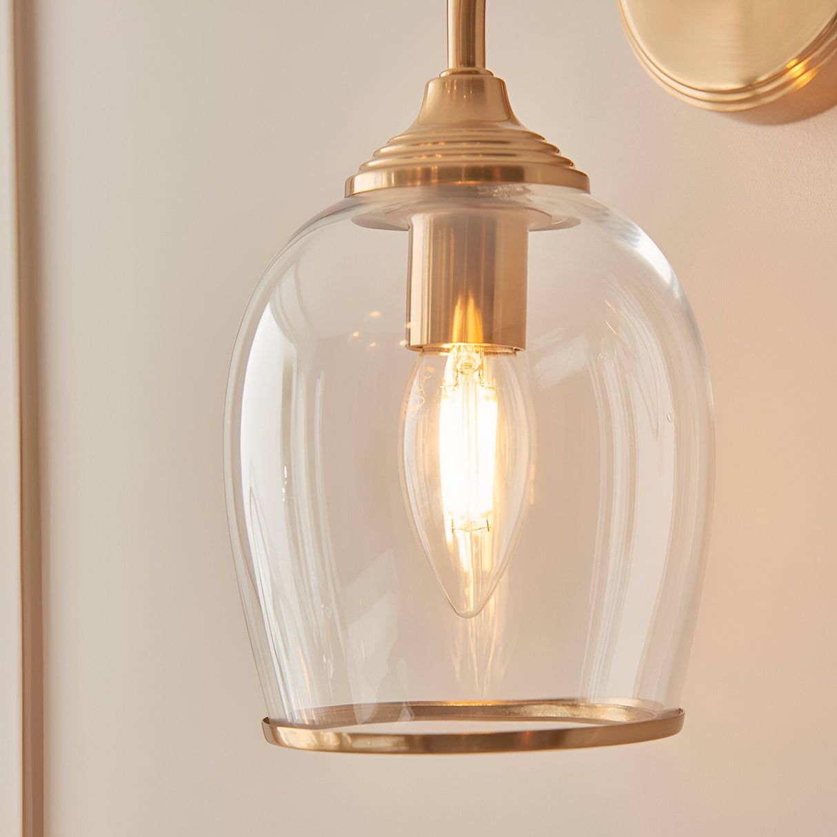 Halstead Wall Light in brass featuring a clear domed glass shade, curved arm, and round back plate, shown lit with a candle-style bulb against a neutral wall.