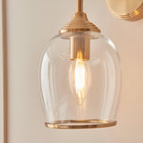 Halstead Wall Light in brass featuring a clear domed glass shade, curved arm, and round back plate, shown lit with a candle-style bulb against a neutral wall.