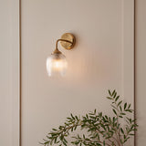 Endon Halstead Ribbed Wall Light in brass with a clear fluted domed glass shade and curved arm, mounted on a neutral panelled wall above olive foliage.