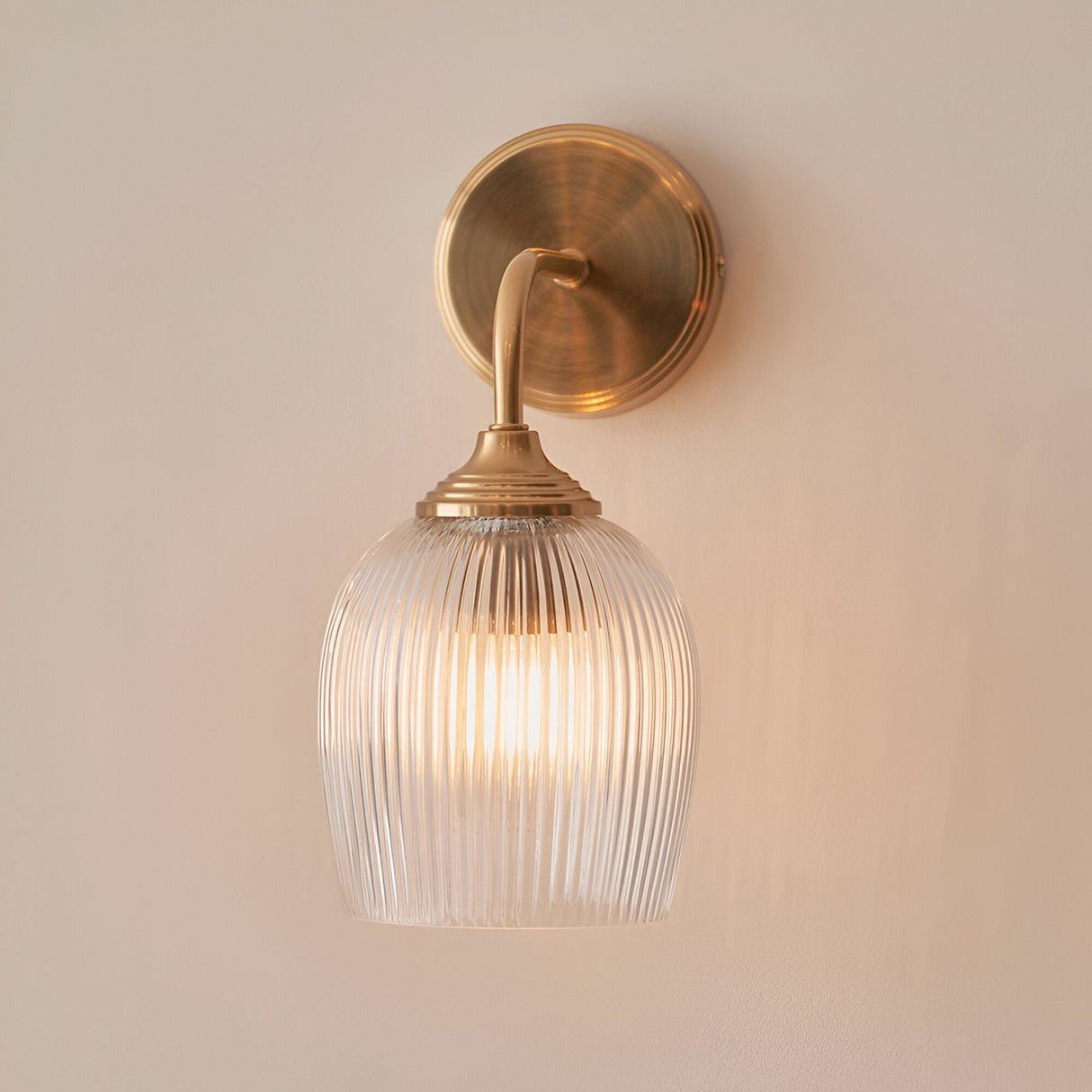 Endon Halstead Ribbed wall light with a clear fluted domed glass shade and brushed brass curved arm and round back plate, mounted on a neutral wall.
