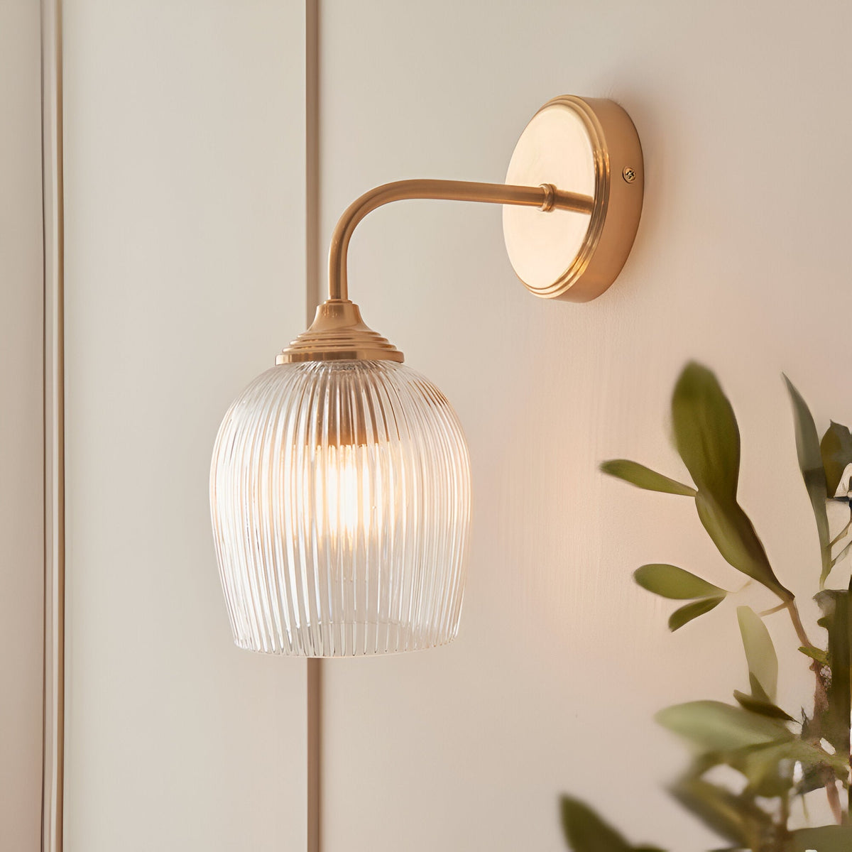 Endon Halstead Ribbed Wall Light in brass with a curved arm, round back plate and clear fluted glass dome shade, mounted beside panelling near foliage.