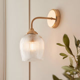 Endon Halstead Ribbed Wall Light in brass with a curved arm, round back plate and clear fluted glass dome shade, mounted beside panelling near foliage.