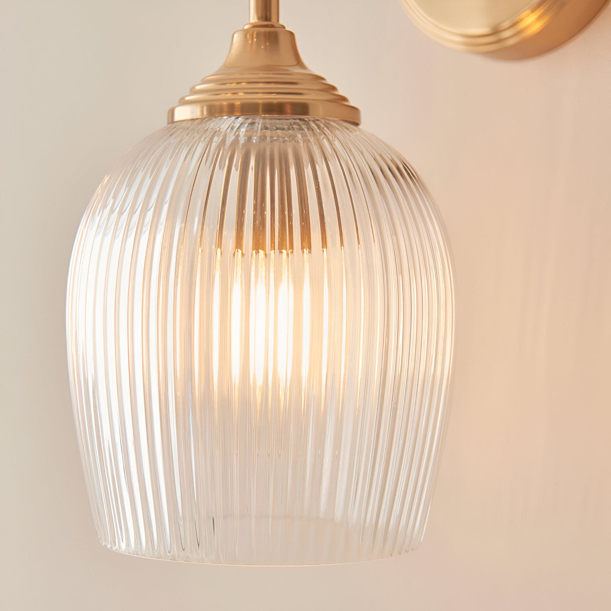 Endon Halstead wall light in brushed brass with a clear ribbed domed glass shade, illuminated close-up against a neutral wall.