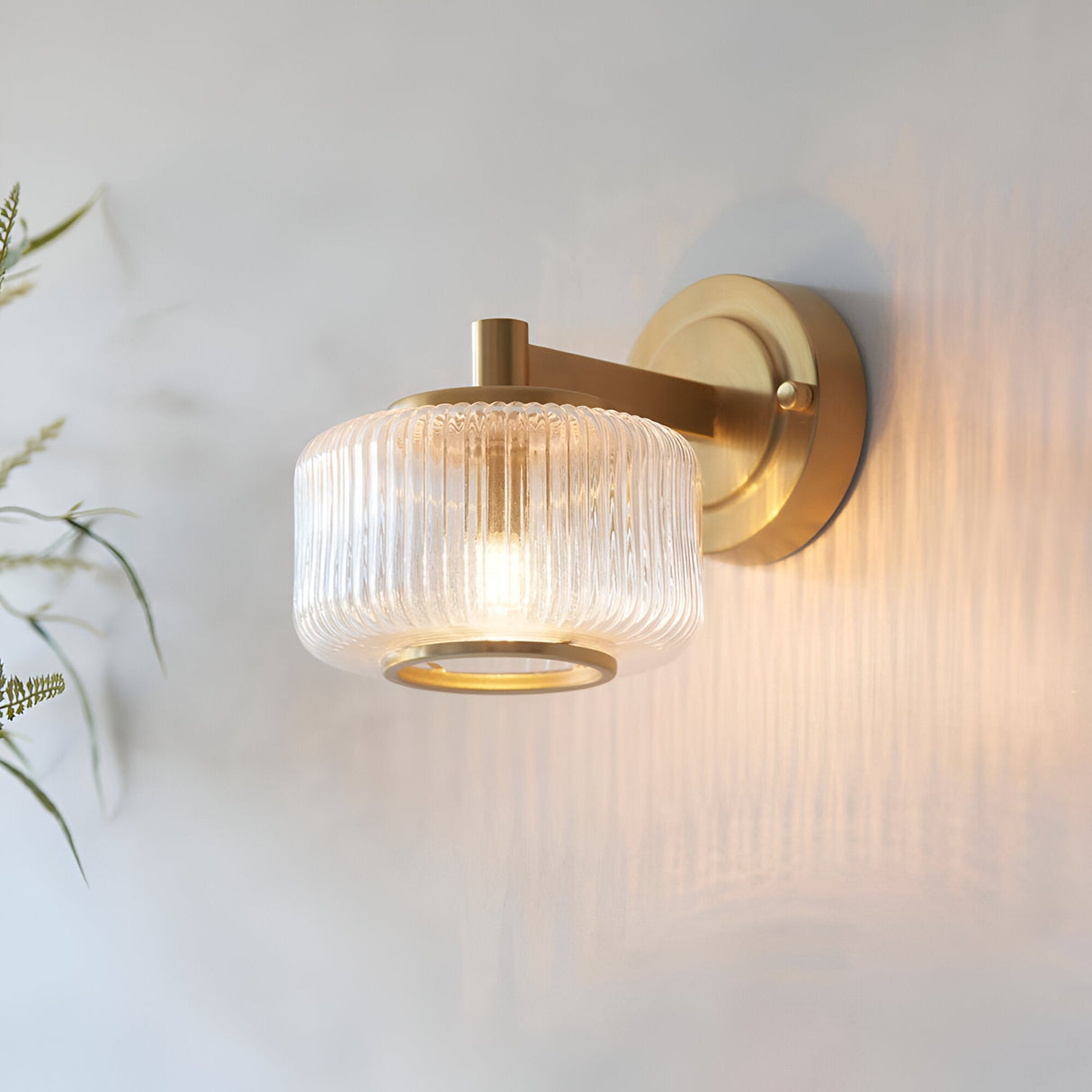 Endon Rutland wall light in brushed aged brass with a ribbed clear glass shade, illuminated on a light wall beside soft green foliage.