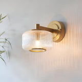 Endon Rutland wall light in brushed aged brass with a ribbed clear glass shade, illuminated on a light wall beside soft green foliage.