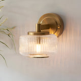 Endon Rutland wall light in brushed aged brass with a ribbed clear glass shade, illuminated on a light wall beside dried grasses.