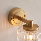 Endon Rutland wall light close-up showing the brushed aged brass circular backplate, knurled screws, arm, and ribbed clear glass shade top.