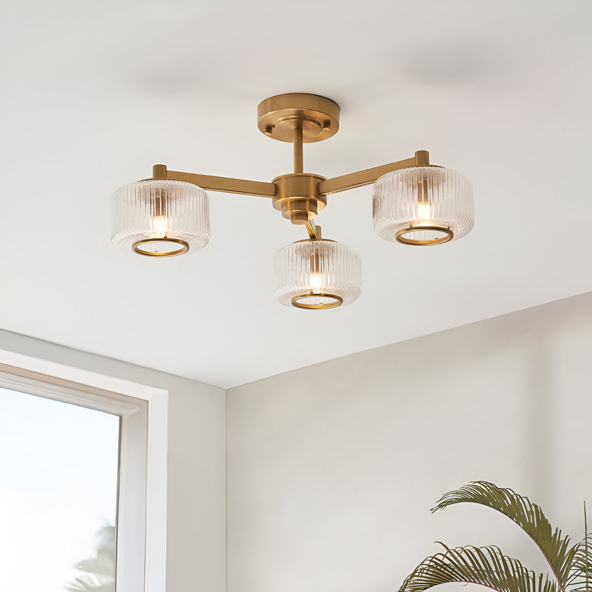 Endon Rutland 3 light semi-flush ceiling light in aged brass with ribbed glass shades, mounted on a white ceiling beside a window and palm plant.