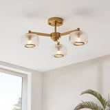 Endon Rutland 3 light semi-flush ceiling light in aged brass with ribbed glass shades, mounted on a white ceiling beside a window and palm plant.