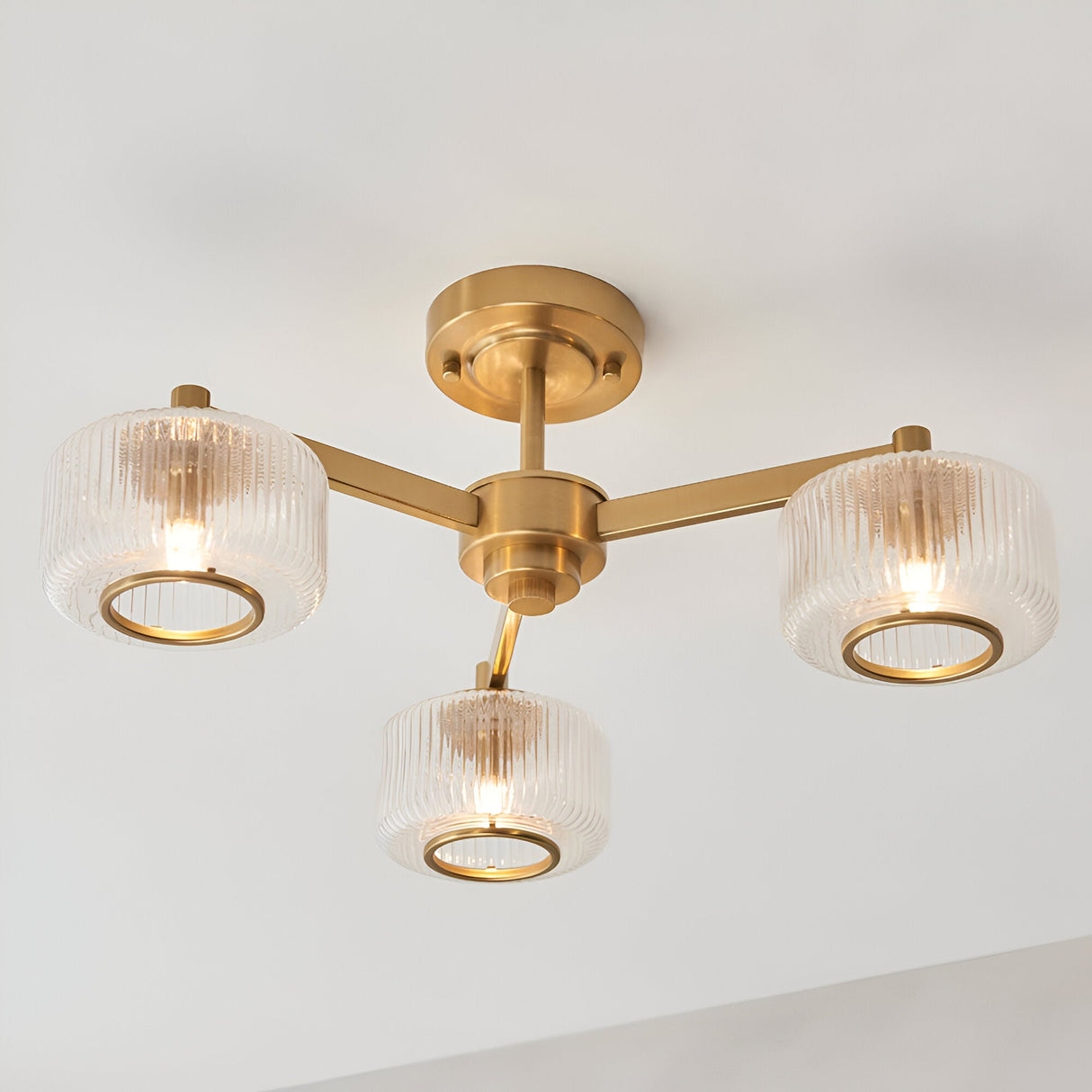 Endon Rutland 3 light semi-flush ceiling light in aged brass with ribbed glass shades on three arms, mounted to a white ceiling.