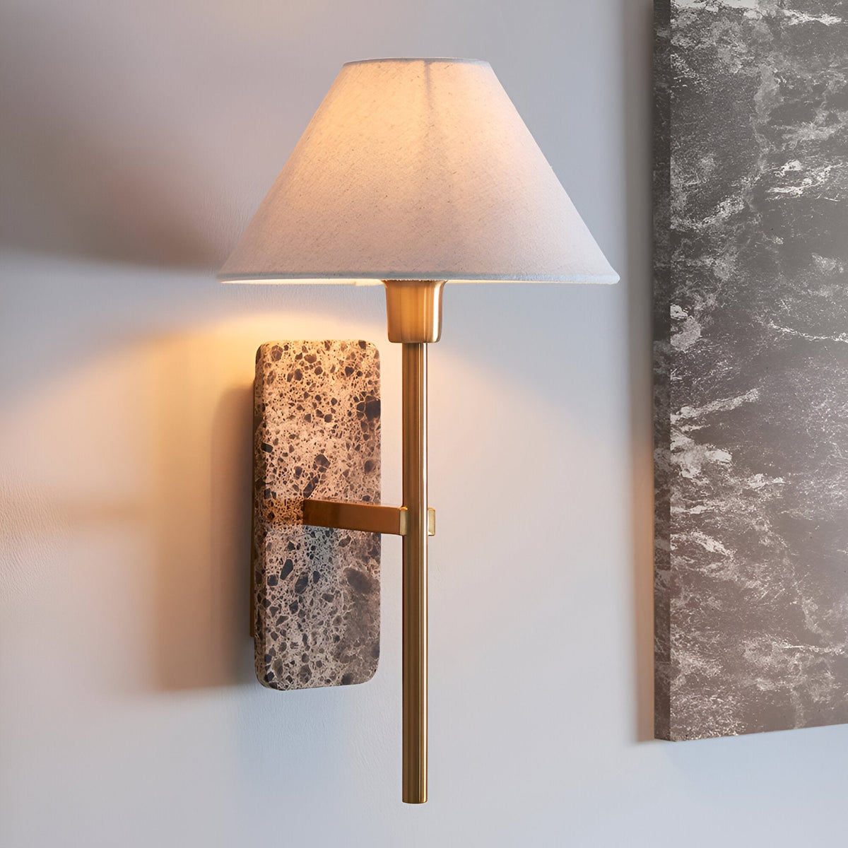 Bramble wall light with a tapered white linen shade, slim brass-toned arm and natural stone backplate, shown illuminated on a light wall beside a marble panel.