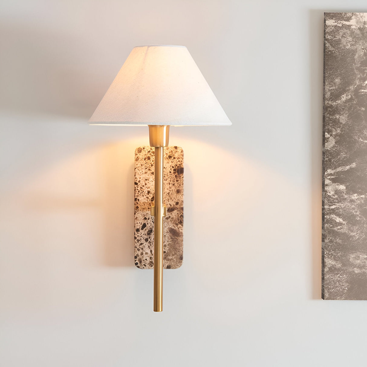 Bramble wall light with a natural stone backplate, slim brass-toned arm and tapered white linen shade, shown lit against a plain wall.
