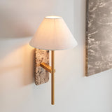 Bramble wall light with natural stone backplate, slim brass-toned arm and tapered white linen shade, mounted beside a marble panel.