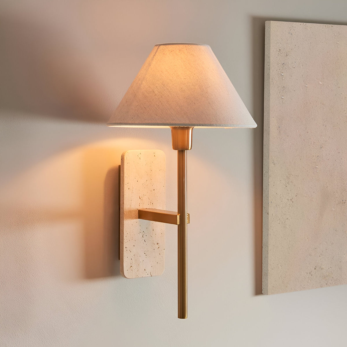 Bramble Wall Light in brass and natural stone, with a tapered linen shade and slim brass arm, shown illuminated on a light wall beside a stone panel.