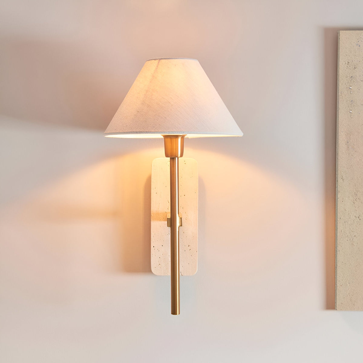 Bramble Wall Light in brass and natural stone, single-light wall fitting with tapered linen shade, mounted on a light wall and glowing warmly.