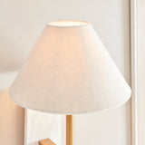 Bramble wall light with a tapered natural linen shade, brass-toned arm and marble backplate, shown lit against a plain light background.