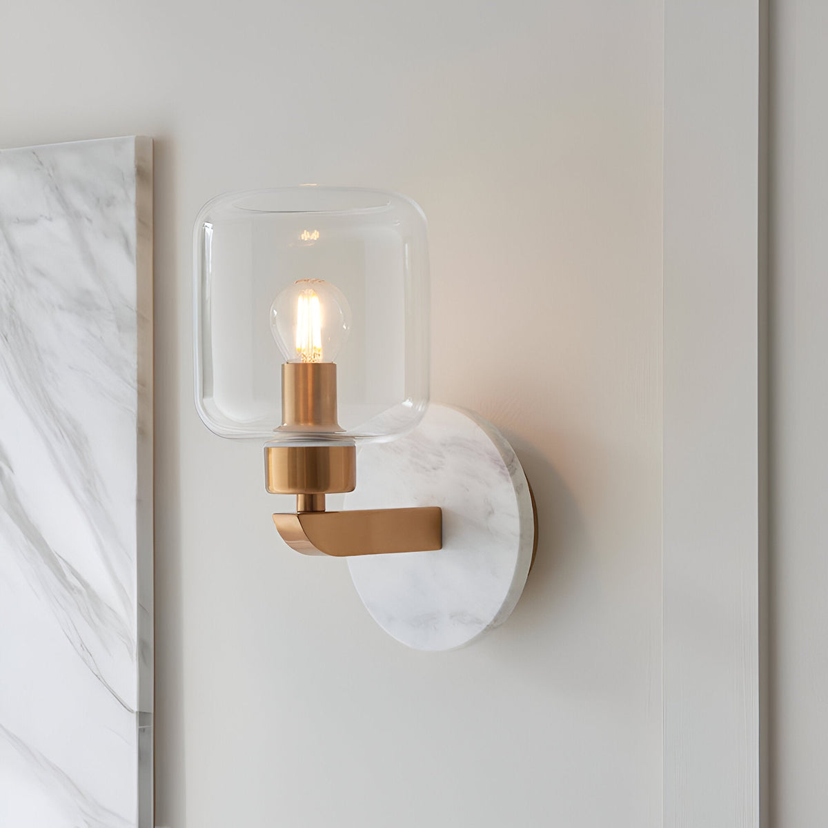 Cranstead Wall Light in brass and white marble, featuring a clear glass shade with visible bulb, mounted on a bedroom wall beside a marble panel.