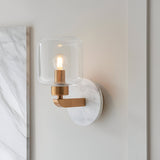 Cranstead Wall Light in brass and white marble, featuring a clear glass shade with visible bulb, mounted on a bedroom wall beside a marble panel.