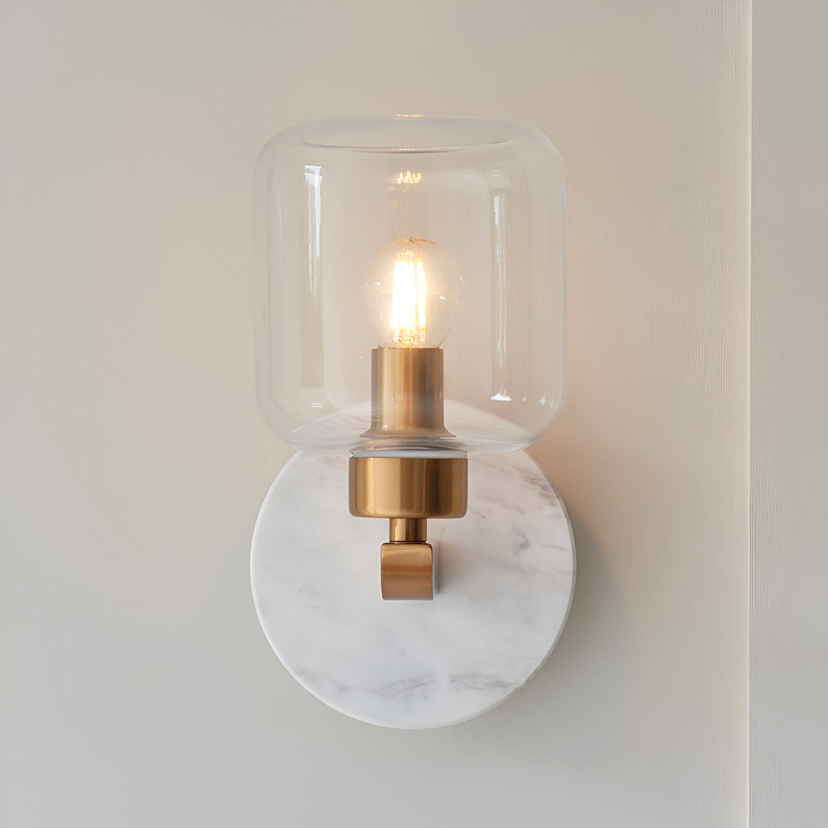 Cranstead Wall Light in Brass & White featuring a round white marble backplate, brass arm, and clear glass shade with a warm E14 bulb, on a plain wall.