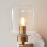 Cranstead Wall Light in brass and white, with a clear glass dome shade over a warm LED bulb, brass arm, and white marble backplate.