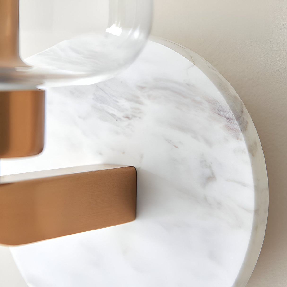 Cranstead Wall Light close-up showing white marble backplate, brass arm and bracket, and curved clear glass shade against a light wall.