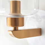 Close-up of the Cranstead Wall Light showing the brushed brass fitting, curved marble backplate, and clear glass shade with visible bulb holder.