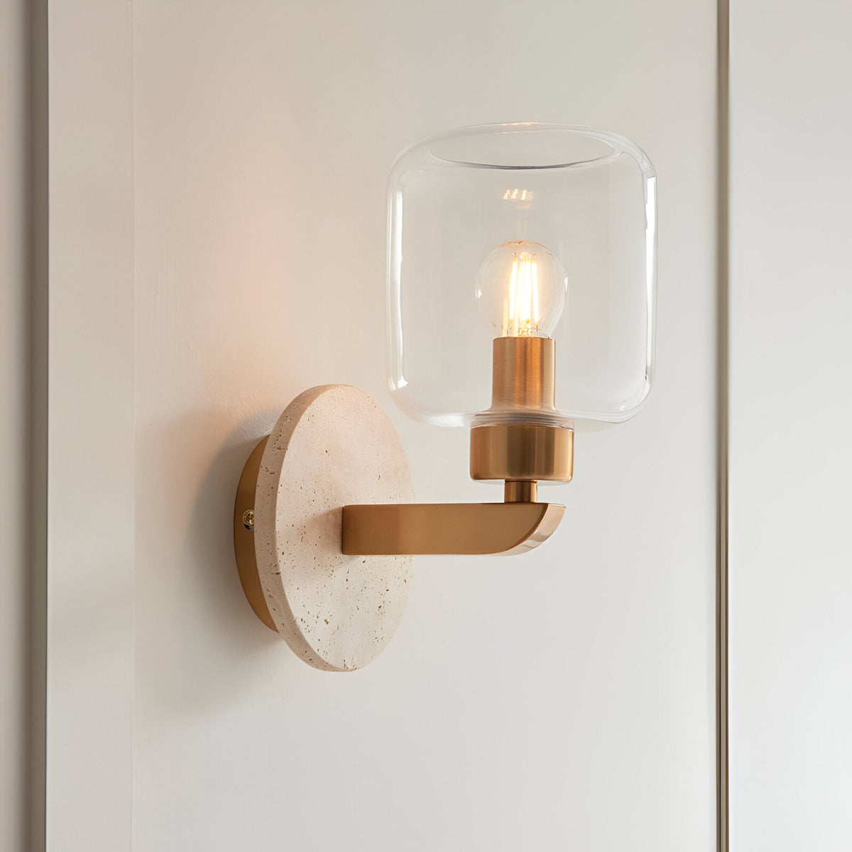 Endon Cranstead wall light with round natural travertine backplate, brass arm and clear glass shade housing a lit E14 filament bulb, on a white wall.