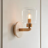 Endon Cranstead wall light with round natural travertine backplate, brass arm and clear glass shade housing a lit E14 filament bulb, on a white wall.