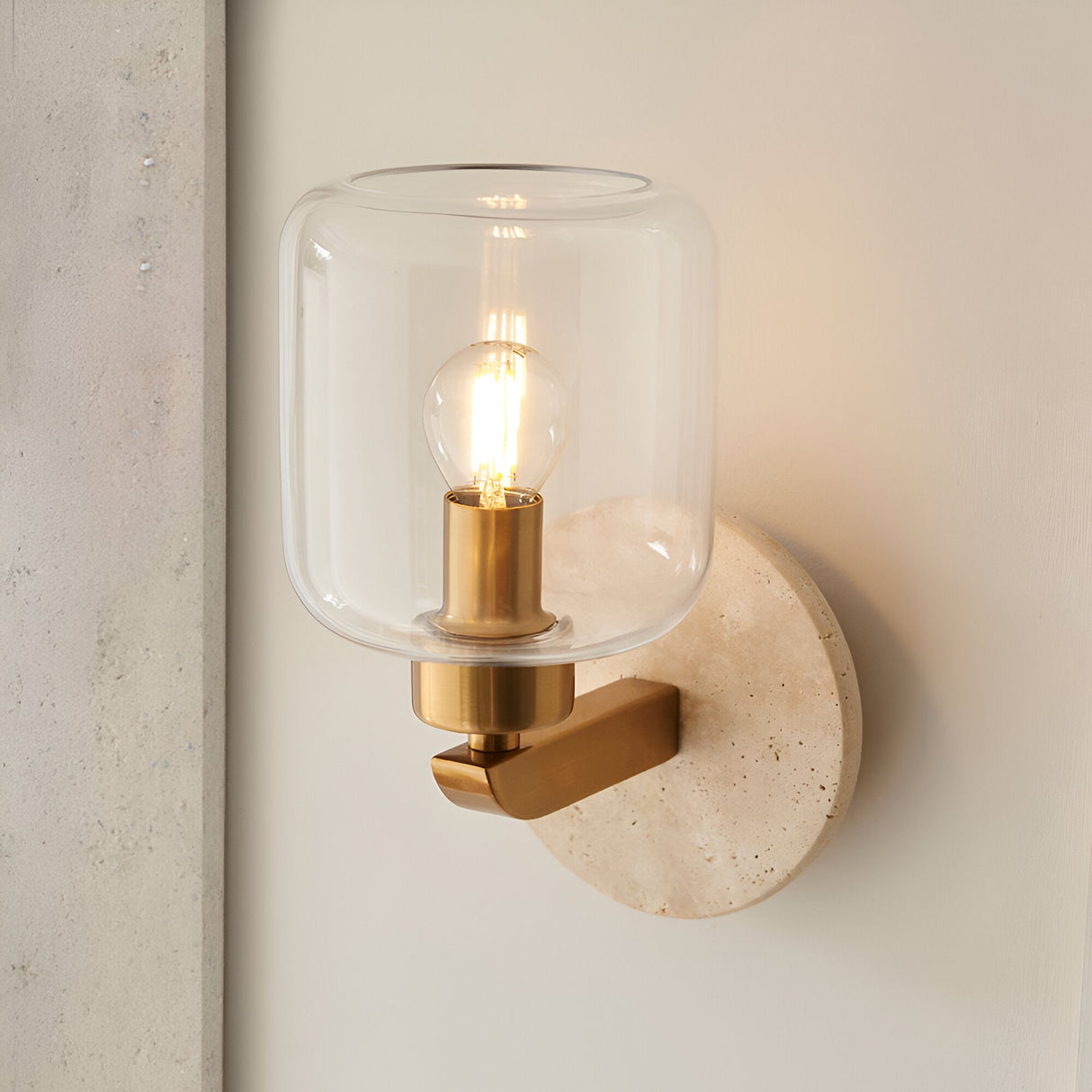 Endon Cranstead wall light with round natural travertine backplate, brass arm, and clear glass shade housing a lit E14 filament bulb.