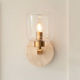 Endon Cranstead wall light with travertine round backplate, brass arm, clear glass shade and lit E14 filament bulb on a neutral wall.