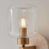 Endon Cranstead wall light close-up showing clear glass cylindrical shade, lit E14 filament bulb and brass fitting on a natural travertine backplate.