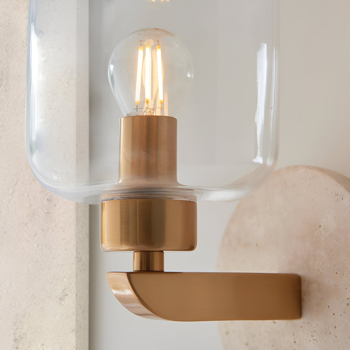 Close-up of Endon Cranstead wall light with brass arm, clear glass shade, E14 filament bulb and natural travertine stone backplate.