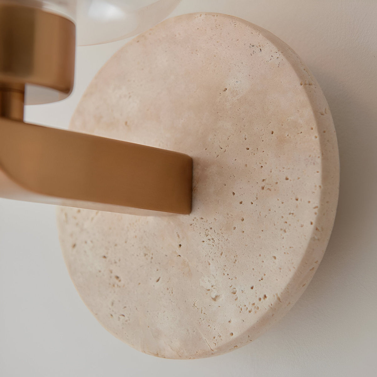 Close-up of Endon Cranstead wall light showing round natural travertine backplate, brushed brass arm, and base of clear glass shade.