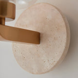 Close-up of Endon Cranstead wall light showing round natural travertine backplate, brushed brass arm, and base of clear glass shade.