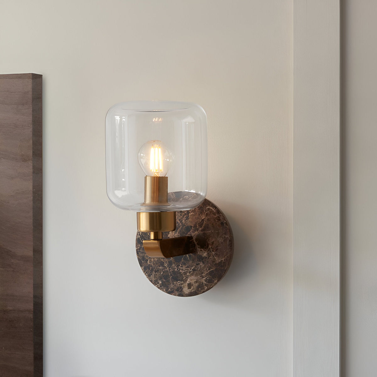 Cranstead wall light with dark brown Emperador marble backplate, brass arm and clear glass shade, mounted on a light wall.