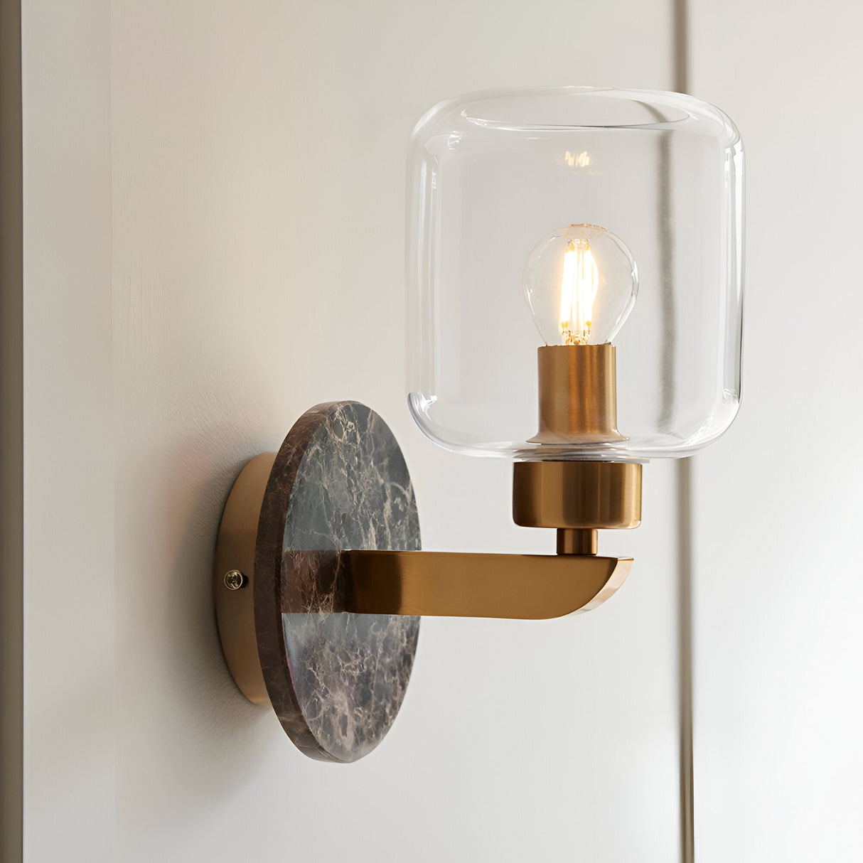 Cranstead Wall Light with brown marble backplate, brass arm and clear glass shade, mounted on a light wall with visible filament bulb.