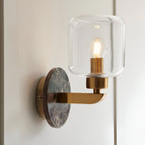 Cranstead Wall Light with brown marble backplate, brass arm and clear glass shade, mounted on a light wall with visible filament bulb.
