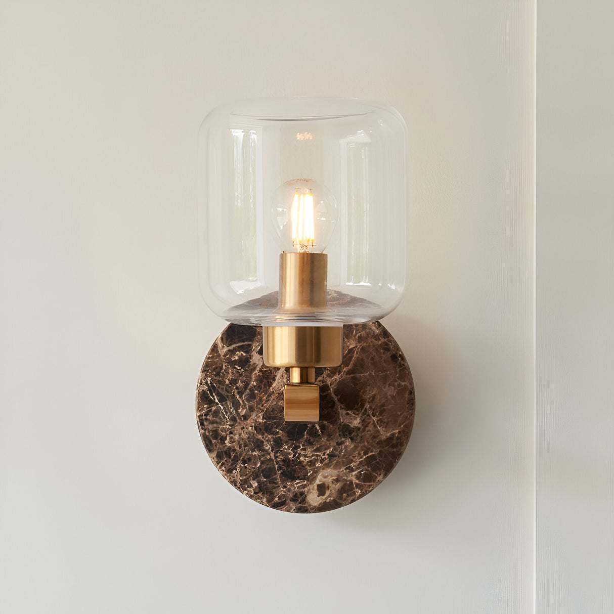 Cranstead Wall Light in Brass & Brown, featuring a round Emperador marble backplate, brass arm, and clear glass shade around a glowing bulb.