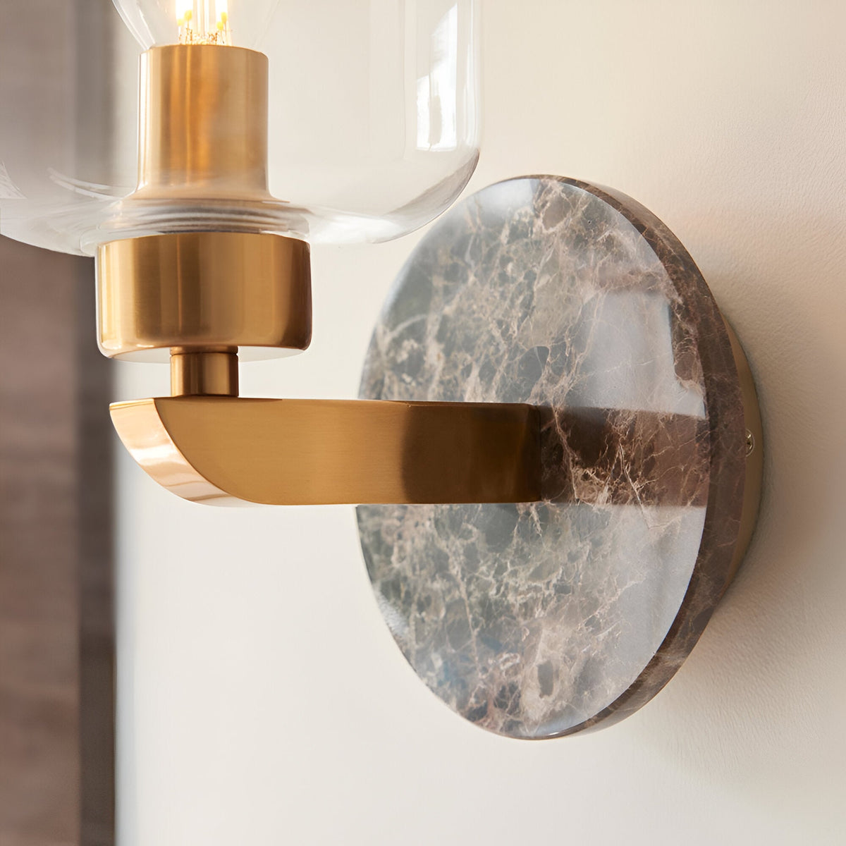 Cranstead wall light with dark brown Emperador marble backplate, brushed brass arm, and clear glass shade revealing the bulb.