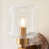 Cranstead Wall Light in Brass & Brown, close-up of clear glass shade with visible filament bulb, brass fitting and dark Emperador marble backplate.