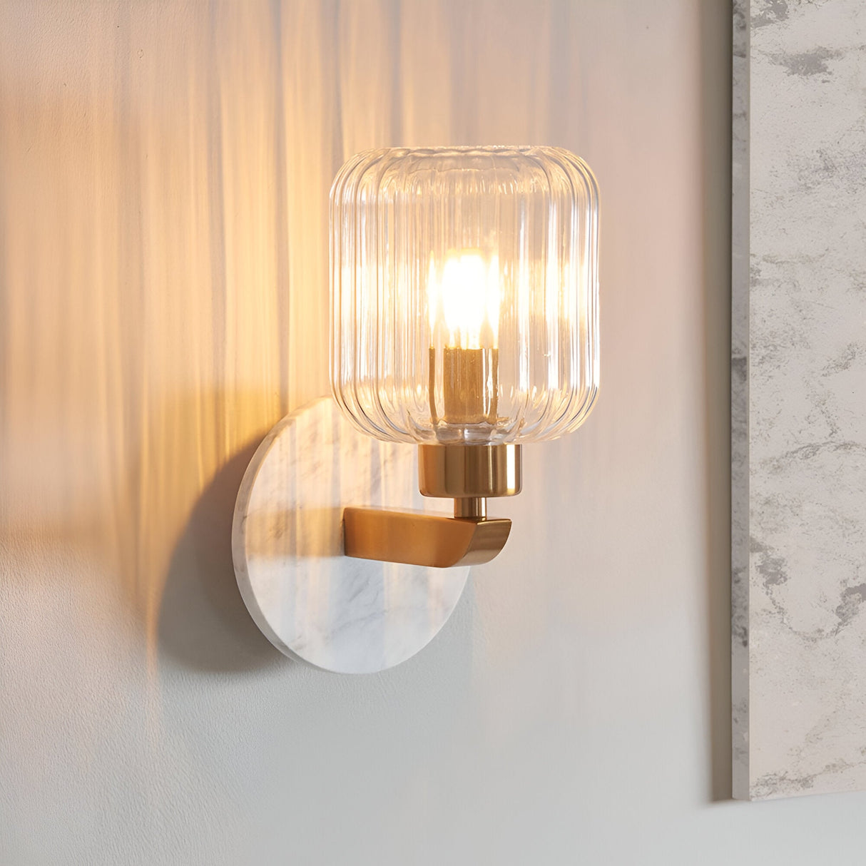 Cranton Wall Light in brass and white, with a round marble backplate, brass arm, and ribbed glass shade lit by an exposed bulb, shown mounted on a wall.