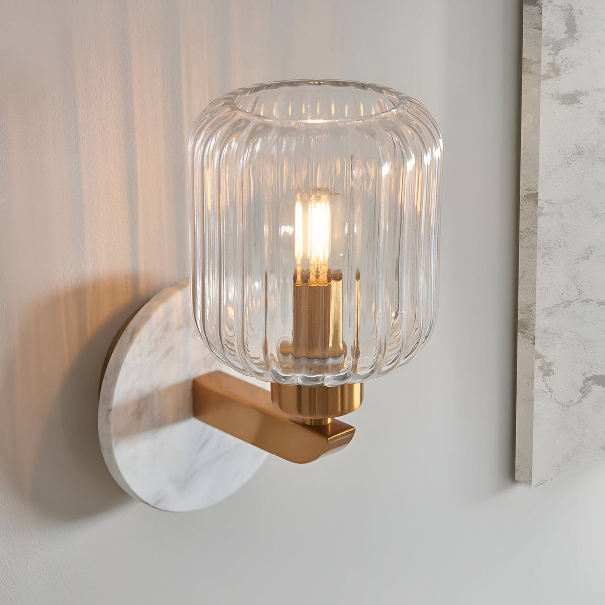 Cranton Wall Light in brass with a white marble backplate and ribbed clear glass shade, shown lit against a plain wall.