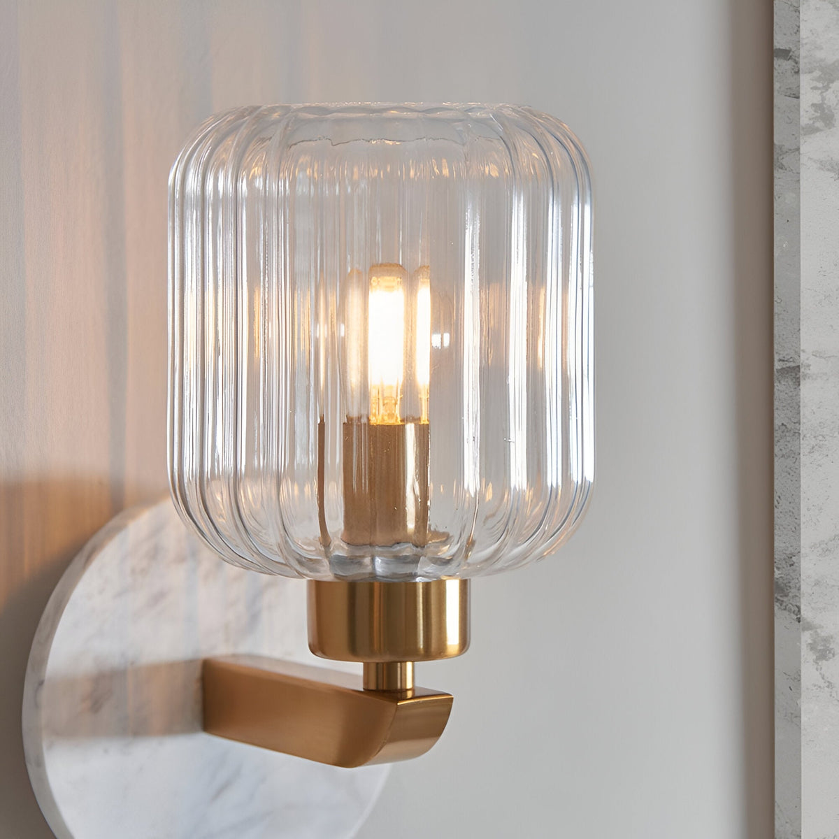 Cranton Wall Light in brass with a ribbed clear glass shade and white marble backplate, shown close-up with a lit bulb against a plain wall.