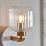 Cranton Wall Light in brass with a ribbed clear glass shade and white marble backplate, shown close-up with a lit bulb against a plain wall.