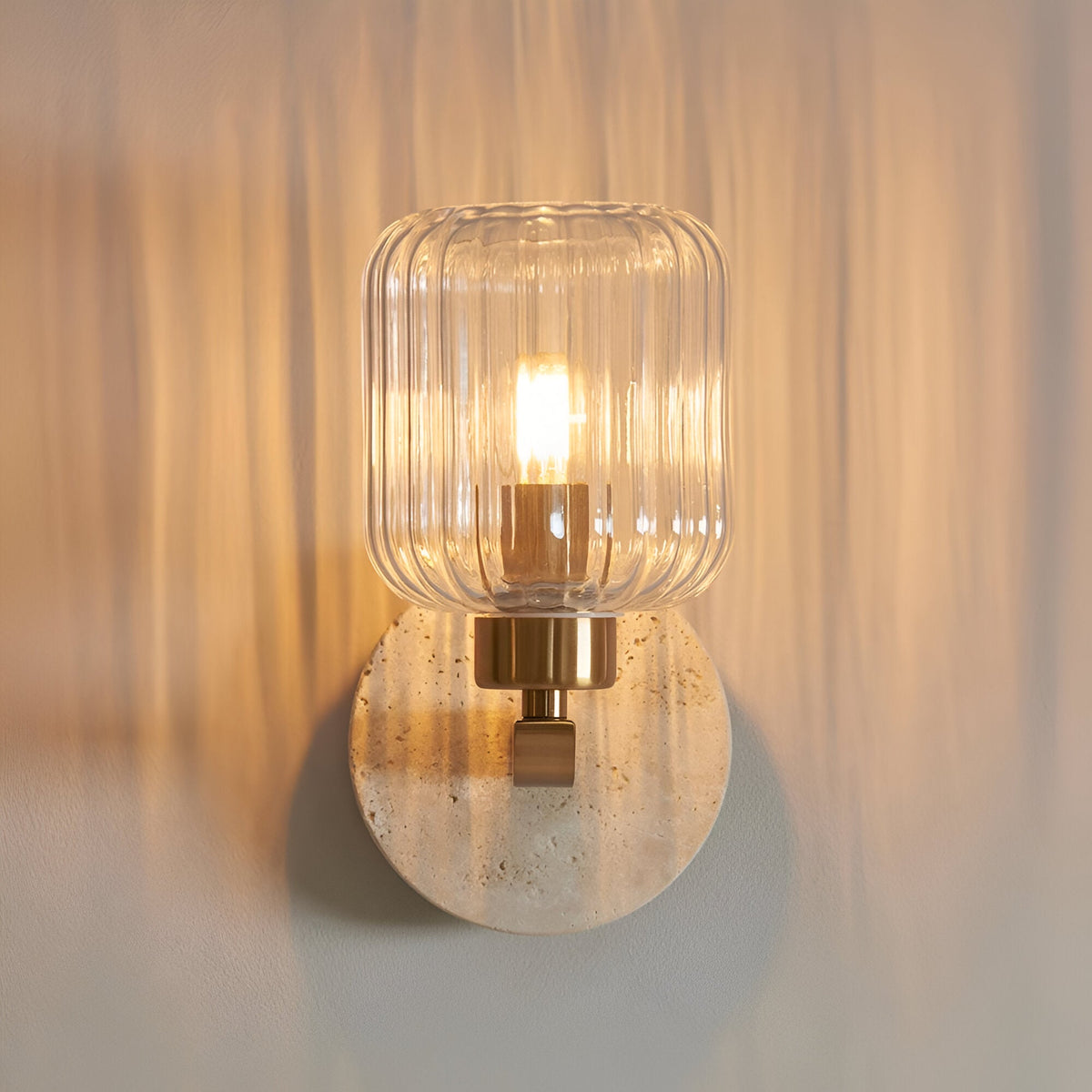 Cranton Wall Light in Brass & Natural, with a round travertine backplate, brass arm and ribbed clear glass shade housing a lit bulb.