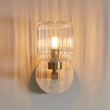 Cranton Wall Light in Brass & Natural, with a round travertine backplate, brass arm and ribbed clear glass shade housing a lit bulb.