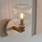 Cranton Wall Light in brass with a ribbed clear glass shade and natural stone backplate, shown close-up with a warm bulb glowing against a light wall.