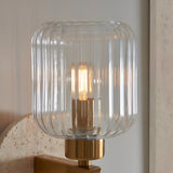 Cranton Wall Light in brass with a ribbed clear glass shade lit by a warm bulb, mounted on a natural stone backplate.
