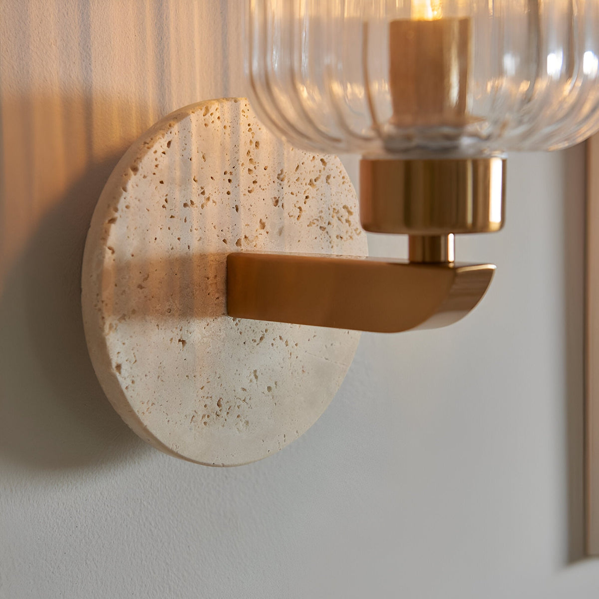 Cranton Wall Light in Brass & Natural, close-up of a round travertine backplate with a brass arm holding a ribbed clear glass shade and visible bulb.
