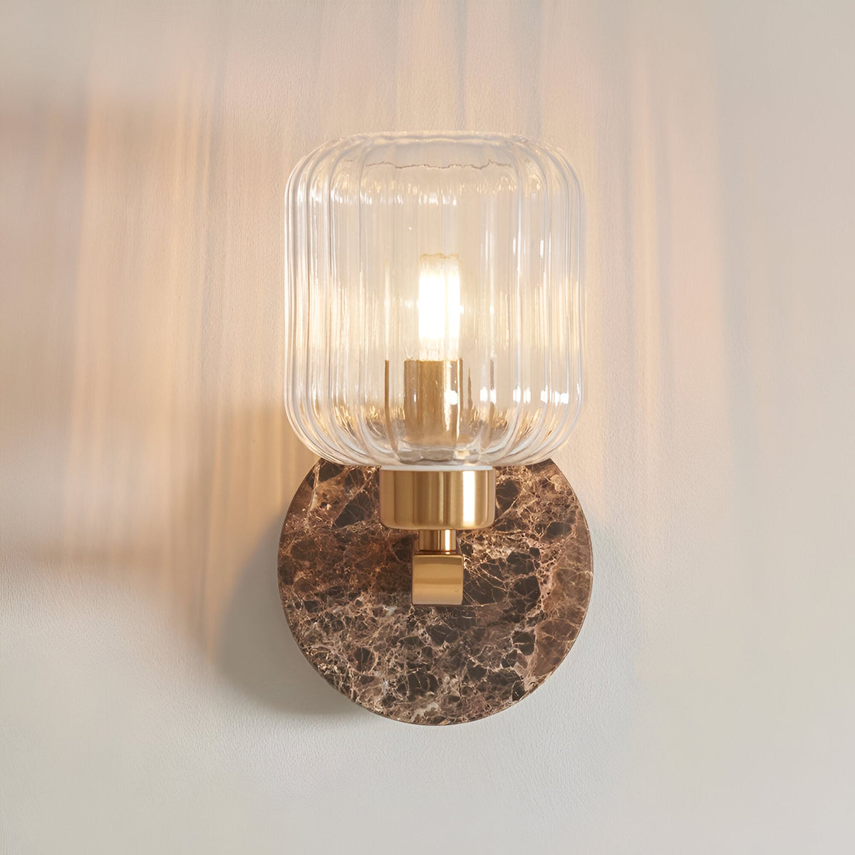 Cranton Wall Light in brass with a round Emperador marble backplate and ribbed clear glass shade, lit, against a plain light background.
